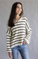 A woman wearing a cream knit button-up shirt with dark brown horizontal stripes and blue jeans, standing against a neutral gray studio background.