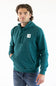 A man wearing a teal green pullover hoodie with a white square The North Face logo patch on the left chest, paired with blue jeans, photographed against a plain white studio background.