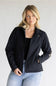 A blonde woman wearing a black zip-up jacket with a small shield logo on the left chest and gold stripes on the sleeve cuffs, paired with a black top and light blue jeans, against a light gray studio background.
