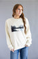 A woman wearing a cream-colored crewneck sweatshirt with a black and white landscape graphic on the front, paired with blue jeans, standing against a solid gray studio background.