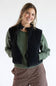 A woman wearing a black zip-up puffer vest over an olive green knit sweater and brown striped pants, photographed against a clean white studio background.