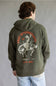 A person viewed from behind wearing an olive green hoodie with a large white and orange graphic on the back, paired with light blue jeans, against a neutral gray studio background.