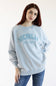 A woman with red hair wearing a light blue washed sweatshirt with 'MICHIGAN GREAT LAKES EST. 1977' printed in blue on the front, paired with blue jeans, against a plain white background.