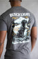 A man viewed from behind wearing a heather gray t-shirt with a large graphic print on the back. The graphic features the text 'BUSCH LIGHT' at the top and 'Head for the Mountains' at the bottom, surrounding an illustration of a black Labrador retriever sitting by a lake with a mountain in the background.