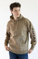 A man wearing a tan pullover hoodie with 'ARIAT' embroidered vertically in brown on the right sleeve and a small yellow logo patch near the cuff, paired with blue jeans against a plain white background.