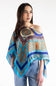 A woman modeling a vibrant turquoise and blue patterned poncho-style top with gold and brown ornate motifs, paired with blue jeans against a plain white studio background.