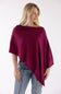 A woman wearing a draped, deep burgundy poncho-style top and light blue jeans, photographed against a plain white studio background.