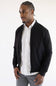 A man wearing a black bomber jacket over a white button-down shirt and gray trousers, photographed against a plain white studio background.