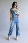 A woman modeling light-wash blue denim overalls with a green short-sleeve top and beige ankle boots, standing against a plain light gray studio background.