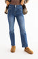 A person wearing medium-wash blue denim jeans with a brown top, a plaid shirt draped off the shoulders, and tan slip-on shoes, photographed against a plain white studio background.