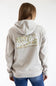 A woman viewed from behind wearing a light beige hoodie with a large back graphic that reads 'Local Girl Outfitters' over a green and beige patterned rectangle, paired with blue jeans against a plain white background.