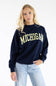 A blonde woman wearing a navy blue crewneck sweatshirt with 'MICHIGAN' printed in yellow across the chest, paired with light blue jeans, against a plain white background.