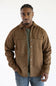 A man wearing a brown button-up overshirt over a green t-shirt and blue jeans, standing against a plain white studio background.