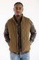 A man wearing a brown diamond-quilted zip-up vest over a camouflage long-sleeve shirt and blue jeans, standing against a plain white background.