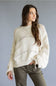 A woman wearing a cream-colored, open-knit cropped sweater with tie details on the cuffs, paired with olive green pants, set against a solid gray studio background.