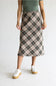 A model wearing a knee-length plaid midi skirt in beige, brown, and black, paired with a sage green top and white platform sneakers, against a plain light gray background.