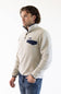 A man wearing a light beige fleece pullover sweater with a navy blue chest pocket and trim, paired with blue jeans, photographed against a plain white studio background.