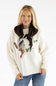 A woman wearing a cream knit sweater with a brown faux-fur collar and a large brown, white, and green graphic on the front, paired with blue jeans, against a plain white background.