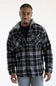 A man wearing a black and gray plaid hooded flannel shirt over a black t-shirt and gray pants, standing against a plain white studio background.