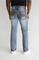 Rear view of a person wearing light-wash blue jeans with decorative brown contrast stitching on the back pockets, paired with a black top and white sneakers, against a light gray studio background.