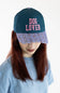 A person wearing a dark teal baseball cap with 'DOG LOVER' embroidered in pink on the front and a patterned brim, paired with a light blue t-shirt against a plain white background.