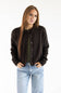 A woman with long light brown hair stands against a plain white studio background wearing a dark brown button-up cardigan over an olive green top and light blue jeans.