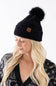 A woman wearing a black plush pom-pom beanie featuring a C.C. branded leather patch on the cuffed hem, paired with a black and white patterned cardigan.
