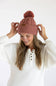 A woman wearing a dusty rose pink cable-knit beanie with a matching pom-pom and a small brown leather brand patch, posing in a white ribbed henley sweater against a clean white background.