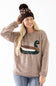 A woman wearing a brown knit beanie with a pom-pom and a taupe sweater featuring a large embroidered duck design on the front, paired with blue jeans, against a plain white background.