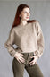 A woman with long red hair wearing a beige knit sweater with raised floral patterns and olive green pants, posing against a neutral gray studio background.