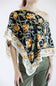 A model wearing a black silk scarf with a yellow rose and green vine pattern, styled as an off-the-shoulder top over olive green pants.