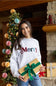 A smiling woman in a white sweatshirt with 'Merry' written across the chest holds a wrapped gift box with a green ribbon. She is standing indoors in front of a decorated Christmas tree and stone fireplace.