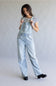 A woman modeling light-wash denim overalls over a black and white striped t-shirt, standing against a neutral gray studio backdrop.