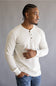 A man wearing a light beige long-sleeve henley shirt and blue jeans, standing against a neutral gray background.