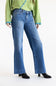 A model wearing a pair of medium-wash blue flare jeans, styled with a light green sweater and black heeled boots against a plain white studio background.