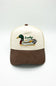A trucker-style baseball cap with an off-white front panel and brim, a brown mesh back and curved brim, and an embroidered mallard duck with the word 'Lucky' on the front.