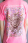 A close-up of a pink short-sleeve t-shirt featuring a large graphic print of a highland cow surrounded by purple flowers, with the text 'BLOOM WITH A KIND HEART' and 'simply southern COLLECTION' on the back.