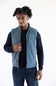 A man wearing a slate blue zip-up vest over a dark navy long-sleeve shirt and blue jeans, standing against a plain white studio background.