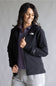 A woman wearing a black The North Face zip-up jacket over a purple collared shirt and gray sweatpants, standing against a neutral gray studio background.