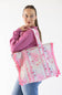 A woman wearing a purple sweatshirt and blue jeans carries a large woven tote bag with pink and white patterns and fringed edges against a plain white background.