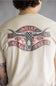 A person shown from the back wearing a cream-colored t-shirt with a large distressed graphic on the back. The graphic features a longhorn skull with an American flag pattern, flanked by banners reading 'ARIAT' and 'ROAM FREE' with '1993' in the center.