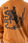 Close-up of the back of a burnt orange sweatshirt featuring a large black graphic print of a cowboy riding a bucking horse, with the word 'ARIAT' and desert illustrations.