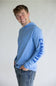 A smiling young man wearing a light blue long-sleeve hooded shirt with 'CARHART' printed vertically in dark blue on the right sleeve, paired with blue jeans, against a plain light gray background.