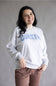 A woman smiling and wearing a light gray crewneck sweatshirt with 'COASTAL' printed across the chest in blue, paired with brown pants, against a plain gray studio background.