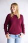 A woman with blonde hair smiling and looking to the side, wearing a textured deep burgundy long-sleeve knit sweater and light blue jeans, standing against a light gray studio background.