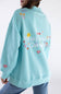 A person wearing an oversized light blue sweatshirt, shown from the back. The sweatshirt features colorful embroidered flowers, hearts, and the phrase 'MAKE HEAVEN CROWded' in white script, with peace signs and small decorative motifs.