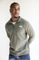 A man wearing an olive green quarter-zip pullover sweater by The North Face, paired with a white t-shirt and blue jeans, photographed against a plain light background.
