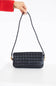 A black woven leather shoulder bag with a braided strap, held by a hand wearing a red sleeve against a clean white background.