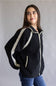 A woman wearing a black sherpa fleece zip-up jacket with cream-colored crochet trim details, paired with blue jeans, photographed against a neutral gray studio background.