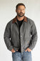 A man wearing a gray waxed cotton utility jacket with a brown corduroy collar, styled over a black t-shirt and blue jeans, standing against a light gray studio background.